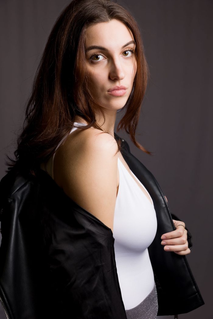 Jeune femme rousse aux yeux verts, vêtue d'un débardeur blanc et d'un perfecto noir, posant en studio avec un regard sensuel