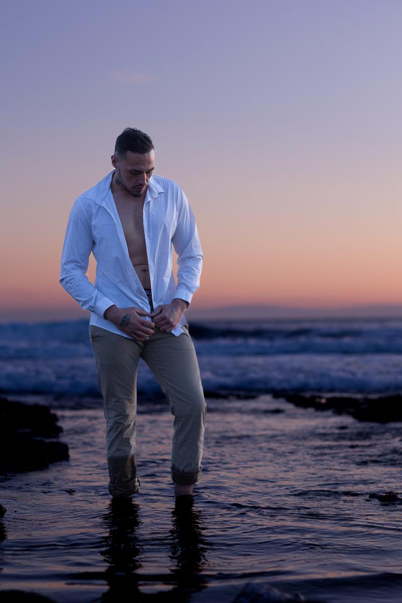 Homme en pantalon beige et chemise blanche ouverte sur ses abdominaux, pieds dans l'eau, photo prise pendant le golden hour à la plage de Carro à Martigues