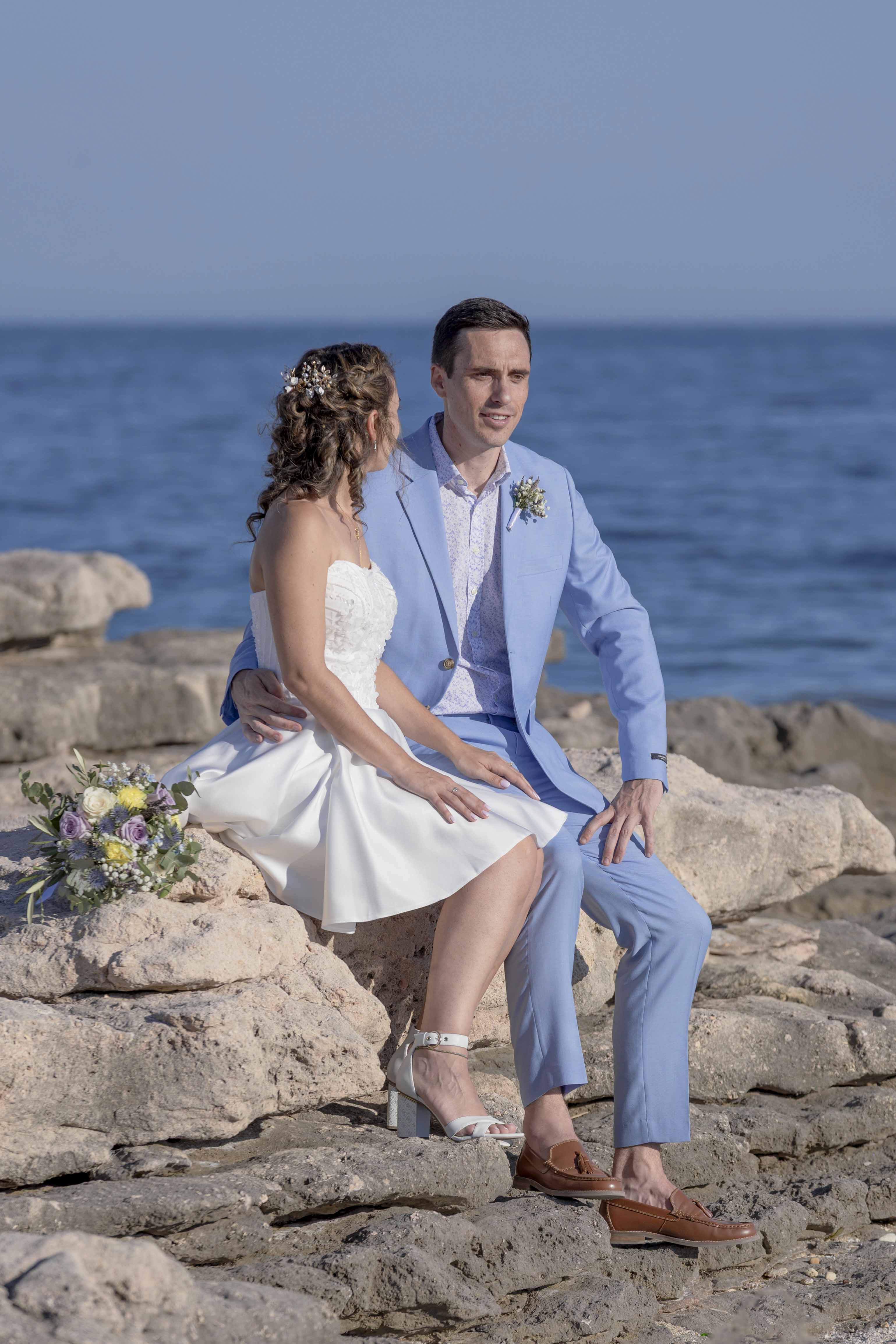 Couple de mariés assis sur les rochers face à la mer, photographie de mariage romantique en bord de mer par M.Studiophoto13