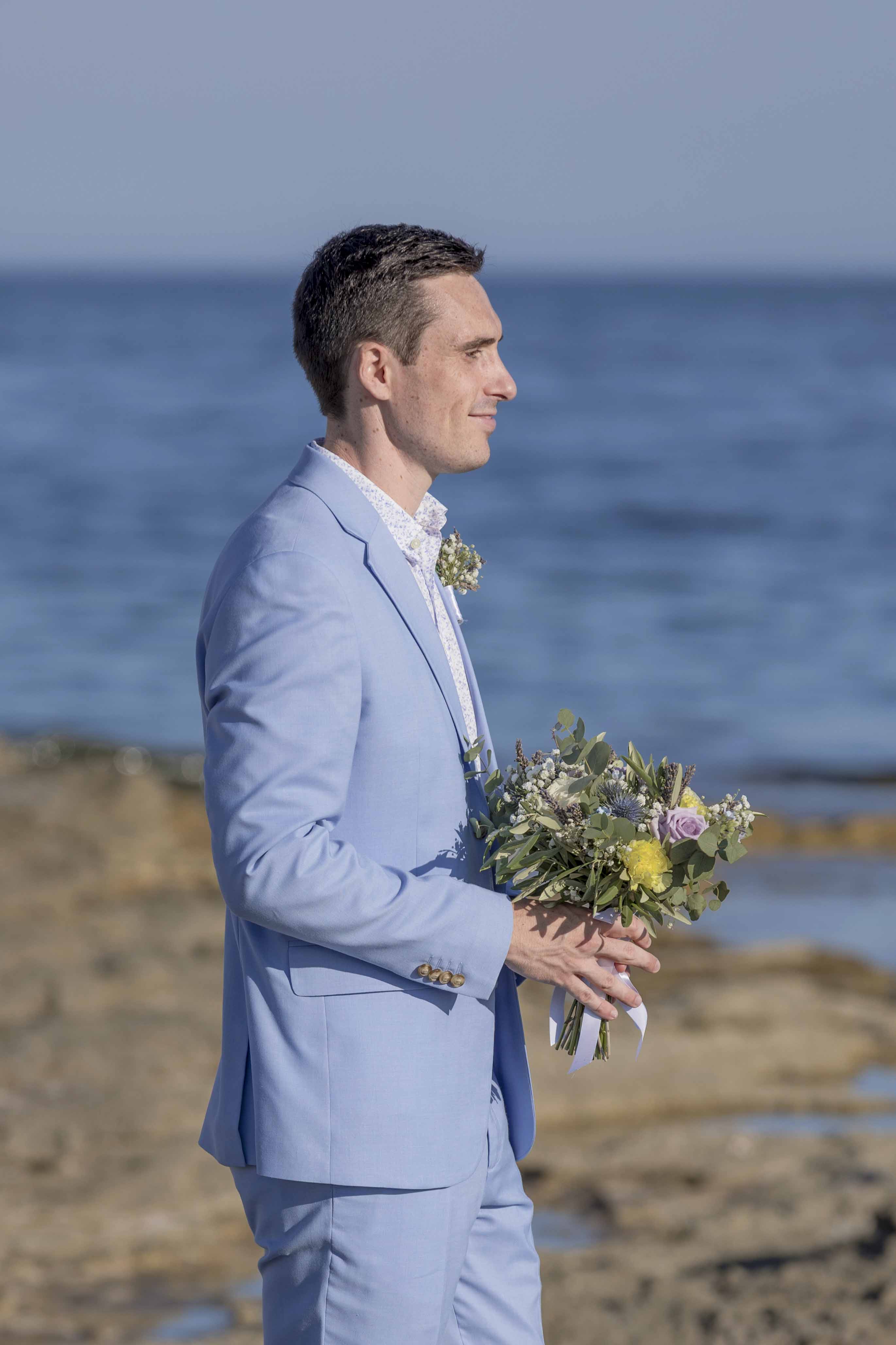 Marié en costume bleu tenant un bouquet de mariage face à la mer – photo par M.Studiophoto13