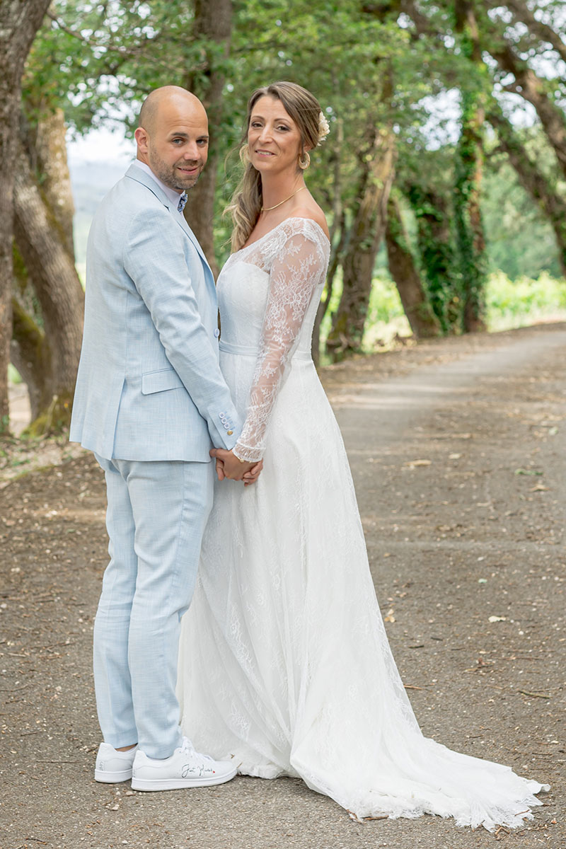 Couple de mariés souriants posant dans une allée bordée d’arbres, photographie de mariage par M.Studiophoto13